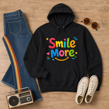 A black pullover hoodie with a colorful 'Smile More' graphic, styled in a flat lay on a wooden surface with blue jeans, white sneakers, sunglasses, and a vintage boombox.