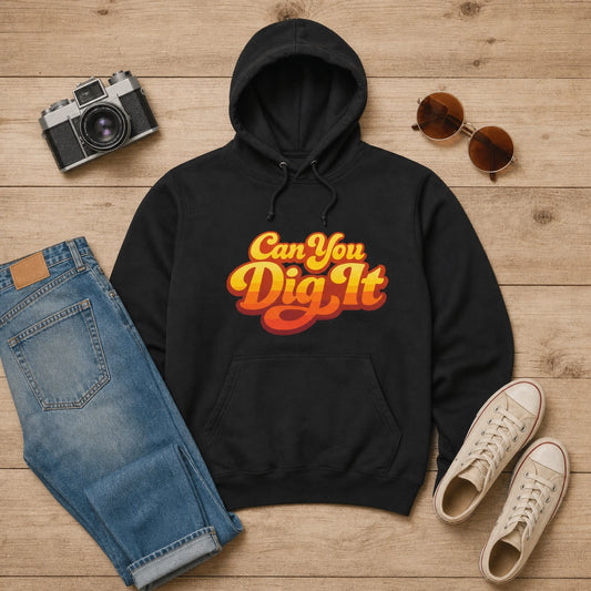 A black pullover hoodie with a retro-style 'Can You Dig It' graphic in orange and yellow, styled in a flat lay on a light wood background with blue jeans, a camera, sunglasses, and white sneakers.