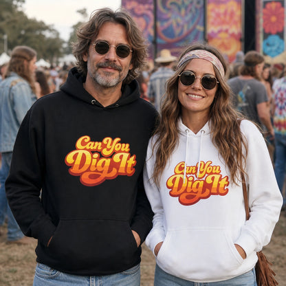 A man and a woman standing outdoors at what looks like a festival or event, each wearing a pullover hoodie with the slogan 'Can You Dig It' in a retro orange and yellow font. The man's hoodie is black, and the woman's is white.