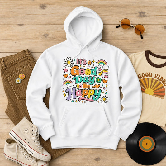 A white pullover hoodie laid flat on a light wood surface, featuring a colorful retro-style graphic that reads 'It's a Good Day to Be Happy' with rainbows, flowers, and hearts. Surrounding props include brown pants, cream sneakers, round sunglasses, a cream graphic T-shirt, and a black vinyl record.