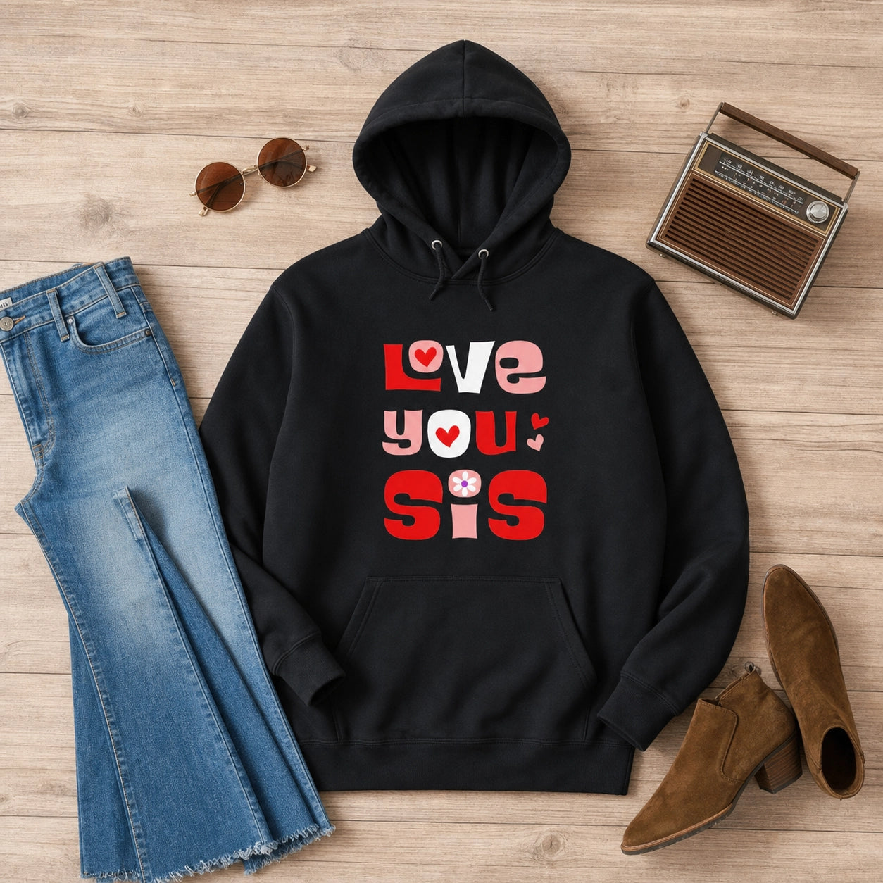 A black pullover hoodie with a colorful 'LOVE YOU Sis' graphic print, styled in a flat lay on a light wood background with blue jeans, sunglasses, a vintage radio, and brown ankle boots.