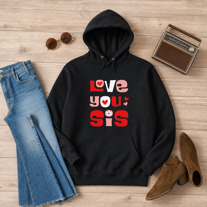 A black pullover hoodie with a colorful 'LOVE YOU Sis' graphic print, styled in a flat lay on a light wood background with blue jeans, sunglasses, a vintage radio, and brown ankle boots.