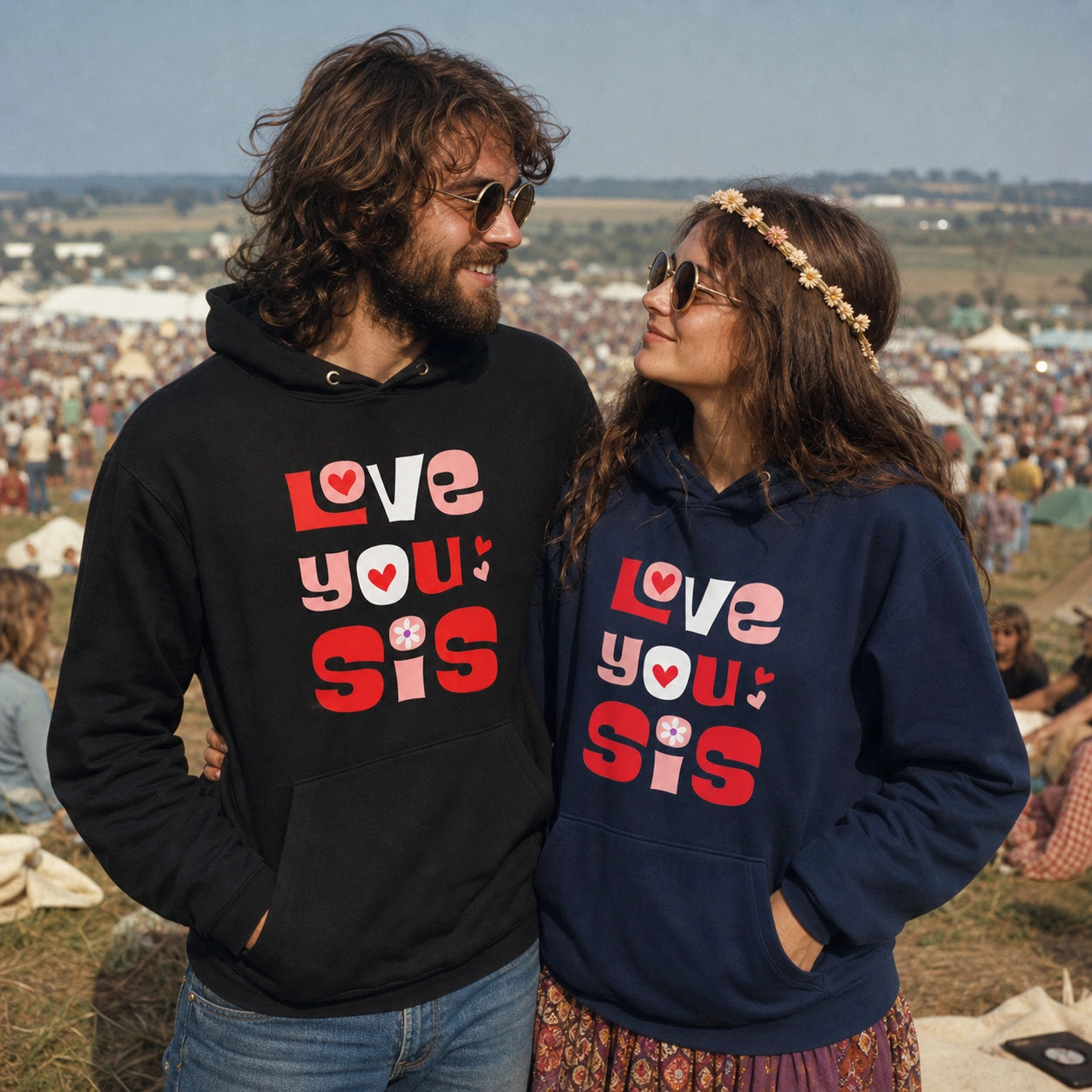 A man and a woman standing outdoors at a large festival, wearing matching hoodies with the text 'Love you sis' printed on the front. The man's hoodie is black, and the woman's hoodie is navy blue.