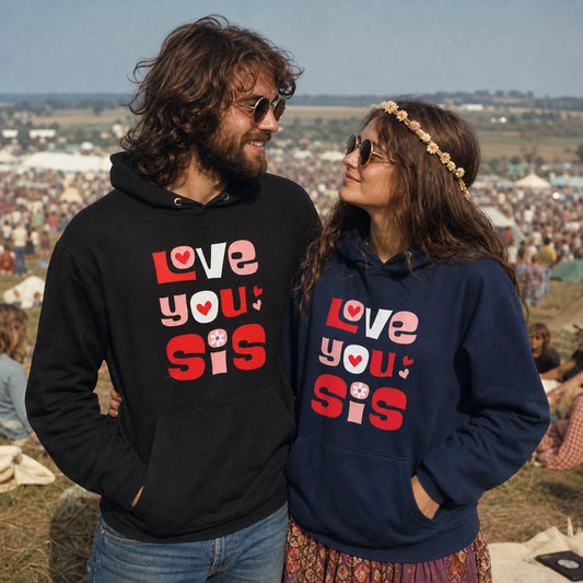 A man and a woman standing outdoors at a large festival, wearing matching hoodies with the text 'Love you sis' printed on the front. The man's hoodie is black, and the woman's hoodie is navy blue.